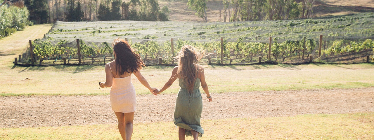 Two girls holding hands walking towards the vines at Stonyridge Vineyard on Waiheke Island