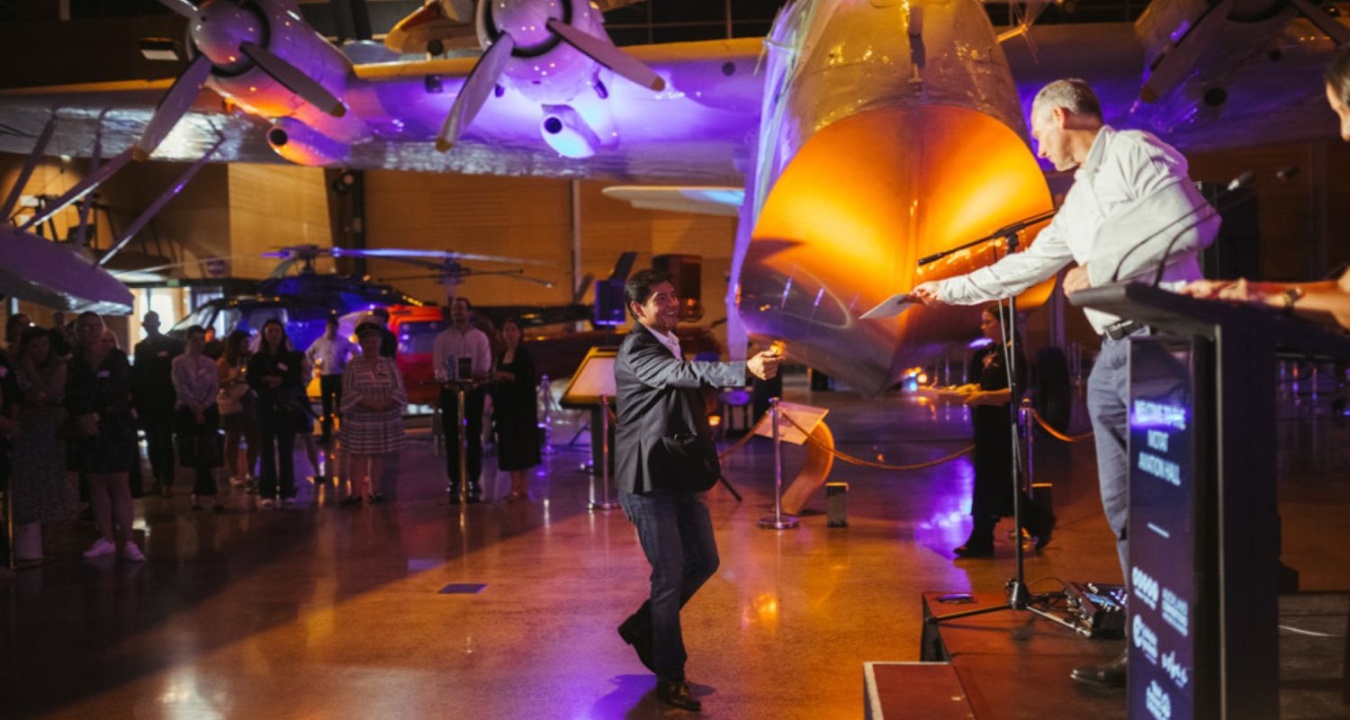 A group watches as a man approaches another man at a podium who is handing him an envelope. They are in the MOTAT aviation hall and there is a plane in the background.